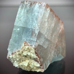 Puede incluir: Una formaci&oacute;n cristalina transl&uacute;cida de color azul claro con una superficie rugosa y texturizada. El cristal tiene forma rectangular con un grupo de peque&ntilde;os cristales beige en su base. Los bordes tienen un tono rojizo-marr&oacute;n.