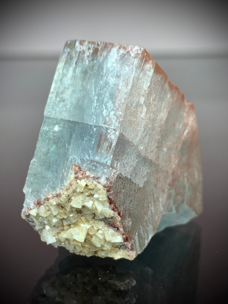 Puede incluir: Una formaci&oacute;n cristalina transl&uacute;cida de color azul claro con una superficie rugosa y texturizada. El cristal tiene forma rectangular con un grupo de peque&ntilde;os cristales beige en su base. Los bordes tienen un tono rojizo-marr&oacute;n.