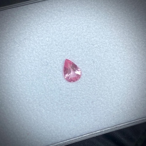 Op de afbeelding: Een peervormige, roze edelsteen. De edelsteen is geplaatst tegen een lichtgrijze achtergrond en heeft een levendige roze tint. De edelsteen is gefacetteerd en reflecteert licht. De presentatie is eenvoudig en elegant, en benadrukt de natuurlijke schoonheid van de edelsteen.