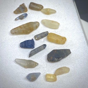 Peut inclure: Une collection de pierres précieuses brutes de différentes formes et couleurs, notamment bleues, jaunes et transparentes. Les pierres sont présentées sur une surface blanche, mettant en valeur leurs textures et facettes naturelles. L'image met en évidence la diversité des pierres.