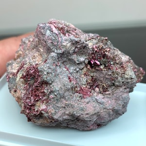 Può includere: Primo piano di una roccia grigia ruvida con vibranti formazioni cristalline color magenta. La roccia ha una superficie strutturata con macchie di tonalità rosa e grigie. L'aspetto generale suggerisce un campione minerale.