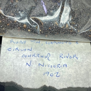 Pode incluir: Um saco plástico transparente cheio de pequenas pedras preciosas escuras. Um rótulo manuscrito diz "Ruby, Tin, Garnets & Zircon, Gurrum River, N. Nigeria, 1907."