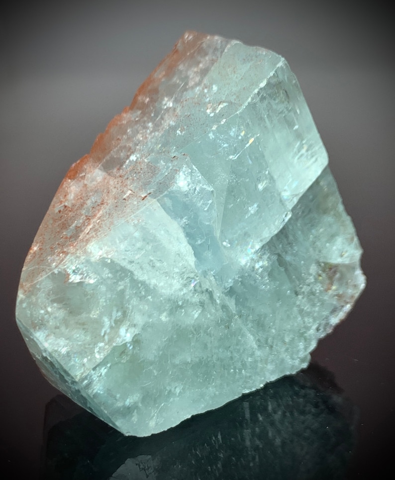 Puede incluir: Una formaci&oacute;n cristalina transl&uacute;cida de color azul claro con una superficie rugosa y texturizada. El cristal tiene forma rectangular con facetas visibles y un toque de coloraci&oacute;n marr&oacute;n rojizo en un borde. La apariencia general es cristalina y natural.