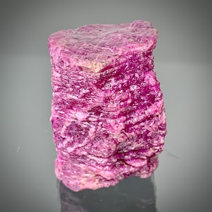 May include: A raw, uncut ruby gemstone with a deep pink hue. The surface has a rough, layered texture, and the stone is cylindrical in shape. The gemstone is displayed against a neutral background, highlighting its natural beauty.