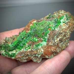 May include: A close-up of a mineral specimen featuring a rough, brown matrix with vibrant green crystalline formations. The green crystals are clustered and textured, contrasting with the earthy tones of the base rock. The specimen is held in a hand.