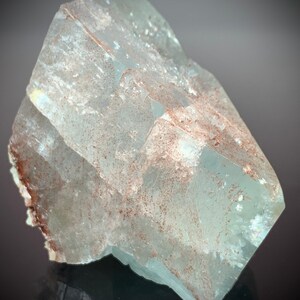 Puede incluir: Una formaci&oacute;n cristalina transl&uacute;cida de color azul claro con forma facetada. El cristal presenta sutiles inclusiones marrones y una superficie reflectante, mostrando la belleza geol&oacute;gica natural. La forma general es angular y geom&eacute;trica, con un aspecto pulido.