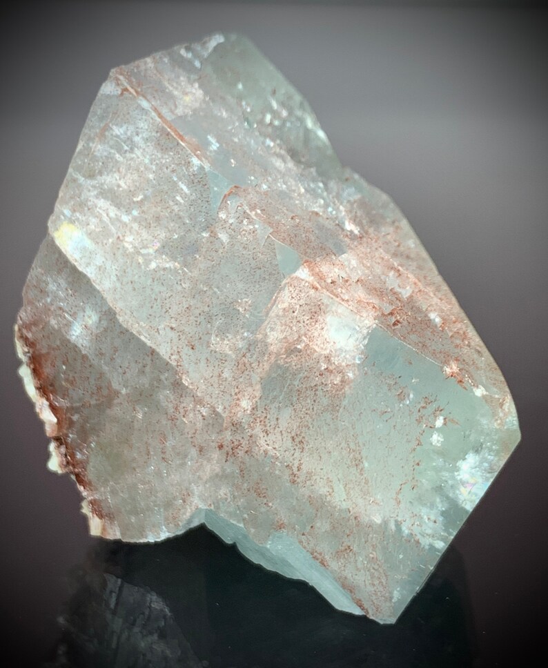 Puede incluir: Una formaci&oacute;n cristalina transl&uacute;cida de color azul claro con forma facetada. El cristal presenta sutiles inclusiones marrones y una superficie reflectante, mostrando la belleza geol&oacute;gica natural. La forma general es angular y geom&eacute;trica, con un aspecto pulido.