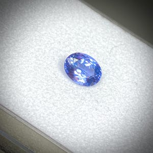 Op de afbeelding: Een ovaalvormige, briljant geslepen edelsteen met een diepblauwe kleur. De edelsteen is geplaatst tegen een witte achtergrond, waardoor de facetten en interne reflecties worden benadrukt. Het gepolijste oppervlak van de steen reflecteert licht.