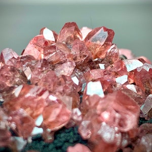 Puede incluir: Primer plano de un grupo de cristales de rodocrosita. Los cristales son de un color rosa-rojizo vibrante, con bordes nítidos y bien definidos. Los cristales están agrupados, creando una exhibición texturizada y llamativa. La base aparece de color verde oscuro.
