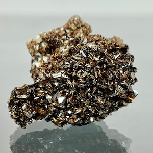 May include: A close-up of a mineral specimen with a cluster of dark brown, metallic-looking crystals. The crystals have a layered, flaky appearance with reflective surfaces, and are set against a lighter brown, rough base. The mineral reflects light.