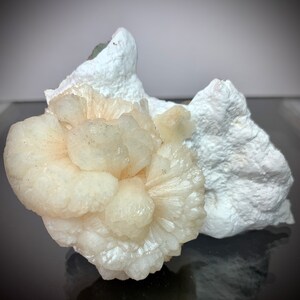 Puede incluir: Un grupo de formaciones cristalinas de color melocot&oacute;n p&aacute;lido con un patr&oacute;n radial, sobre un fondo de roca blanca texturizada. Los cristales tienen una apariencia delicada y en capas, lo que sugiere un esp&eacute;cimen mineral natural.