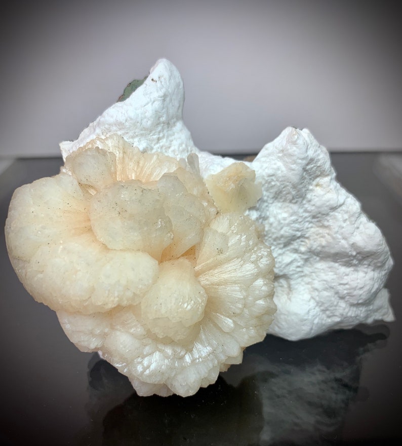 Puede incluir: Un grupo de formaciones cristalinas de color melocot&oacute;n p&aacute;lido con un patr&oacute;n radial, sobre un fondo de roca blanca texturizada. Los cristales tienen una apariencia delicada y en capas, lo que sugiere un esp&eacute;cimen mineral natural.