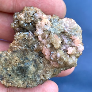以下が含まれることがあります： 鉱物標本のクローズアップ。ベースはくすんだ茶色で、小さな薄ピンク色の結晶のクラスターが表面に散らばっています。質感はざらざらしていて、不均一に見えます。