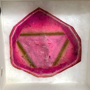 K&ouml;nnte beinhalten: Ein polierter, sechseckiger Edelstein-Schnitt mit einem leuchtend rosa Zentrum, das in einen helleren rosa und wei&szlig;en Rand &uuml;bergeht. Der Stein wird in einer wei&szlig;en Box mit weichem, strukturiertem Futter pr&auml;sentiert, um seine nat&uuml;rliche Sch&ouml;nheit hervorzuheben.