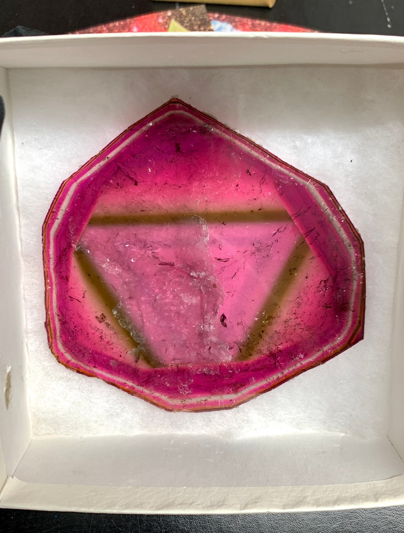 K&ouml;nnte beinhalten: Ein polierter, sechseckiger Edelstein-Schnitt mit einem leuchtend rosa Zentrum, das in einen helleren rosa und wei&szlig;en Rand &uuml;bergeht. Der Stein wird in einer wei&szlig;en Box mit weichem, strukturiertem Futter pr&auml;sentiert, um seine nat&uuml;rliche Sch&ouml;nheit hervorzuheben.