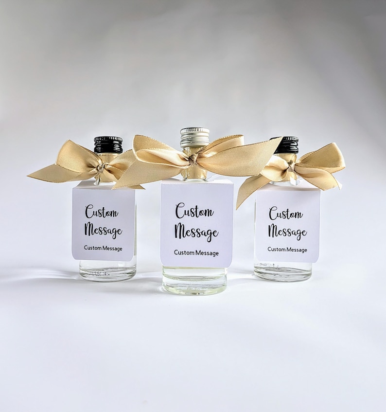 May include: Three small glass bottles with gold ribbon bows and white labels that say "Custom Message" in black text. The bottles are filled with a clear liquid.