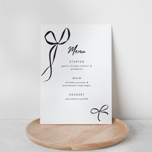 May include: A black and white printable wedding menu with a handwritten font. The menu includes a starter, main course, and dessert. The menu is decorated with hand-drawn bows.