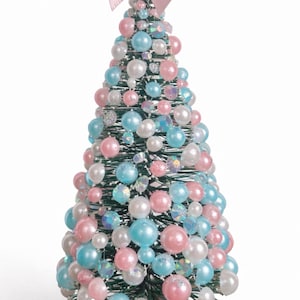 May include: A decorative Christmas tree with a pink bow on top. The tree is adorned with pink, blue, and white beads and iridescent accents. The base is white with glitter.