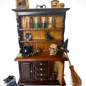 May include: A miniature wooden cabinet with shelves, drawers, and a door, decorated with Halloween-themed items. Includes a skull, a witch's hat, a broom, a black cat, and jars of glitter. A small framed picture and pumpkins are on top.