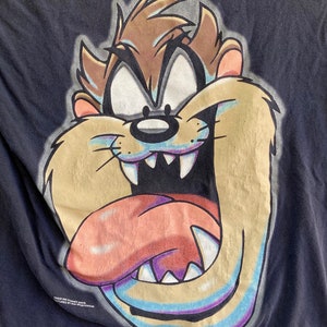 May include: A black t-shirt featuring a large, colourful graphic of Taz the Tasmanian Devil from the Looney Tunes cartoon. Taz is depicted with his mouth open, tongue sticking out, and a menacing expression. The text "Warner Bros. Licensed Product" is printed at the bottom of the shirt.