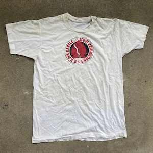 Op de afbeelding: Wit t-shirt met een cirkelvormige afbeelding met een rood en wit ontwerp en de tekst "R. ROE BARTLE SCOUT RESERVATION B.S.A. NOLLIVATION" in een rode en zwarte lettertype. Het shirt is licht gekreukt.