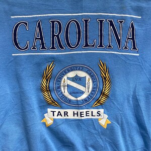 May include: A light blue t-shirt with the word "CAROLINA" in navy blue lettering. Below the word is a gold crest with the words "UNIVERSITAT CAROL. SEPTENT." and "SIGILLVM.  LIBERTAS.  LVX."  The crest is surrounded by two gold laurel wreaths. Below the crest is a white banner with the words "TAR HEELS" in navy blue lettering.