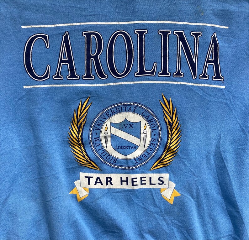 May include: A light blue t-shirt with the word "CAROLINA" in navy blue lettering. Below the word is a gold crest with the words "UNIVERSITAT CAROL. SEPTENT." and "SIGILLVM.  LIBERTAS.  LVX."  The crest is surrounded by two gold laurel wreaths. Below the crest is a white banner with the words "TAR HEELS" in navy blue lettering.