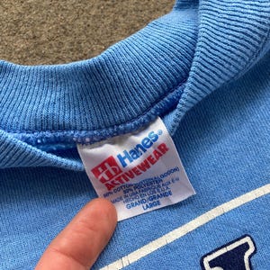 May include: A light blue Hanes Activewear sweatshirt with a white tag that reads "Hanes Activewear 50% Cotton 50% Polyester Made in USA Fabrique aux E.U. Hecho en los E.U. Grand/Grande Large".