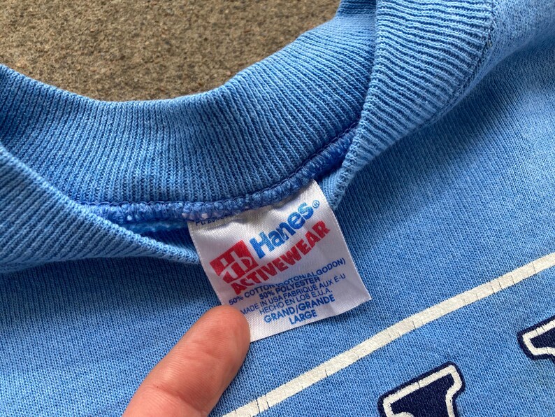 May include: A light blue Hanes Activewear sweatshirt with a white tag that reads "Hanes Activewear 50% Cotton 50% Polyester Made in USA Fabrique aux E.U. Hecho en los E.U. Grand/Grande Large".