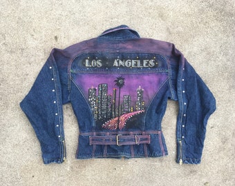 Peut inclure: Une veste en jean bleu foncé avec un motif de ville en violet et noir au dos. Le motif comprend les mots "Los Angeles" et un palmier. La veste a une ceinture et des poches zippées.