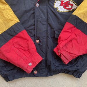 May include: A vintage black, red, and gold jacket with the Kansas City Chiefs logo on the upper chest. The jacket features a snap-button closure and color-blocked sleeves. The sleeves are red, black, and gold.