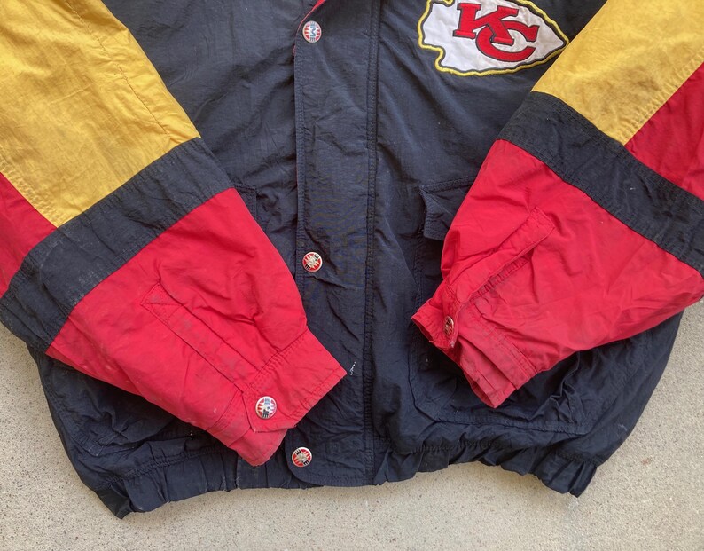 May include: A vintage black, red, and gold jacket with the Kansas City Chiefs logo on the upper chest. The jacket features a snap-button closure and color-blocked sleeves. The sleeves are red, black, and gold.