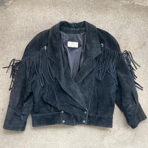 May include: A black suede jacket with fringe detailing on the sleeves and collar. The jacket has a zipper closure and a button-up front.