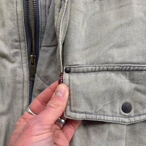 Puede incluir: Primer plano de una chaqueta vaquera gris claro con cierre de cremallera y un peque&ntilde;o bolsillo en el lado izquierdo. El bolsillo tiene una etiqueta roja de Levi's.