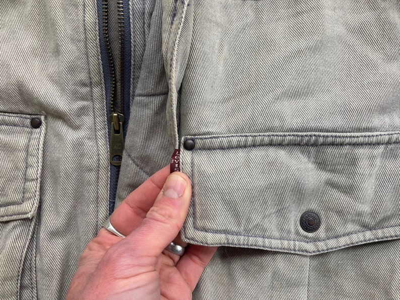 Puede incluir: Primer plano de una chaqueta vaquera gris claro con cierre de cremallera y un peque&ntilde;o bolsillo en el lado izquierdo. El bolsillo tiene una etiqueta roja de Levi's.