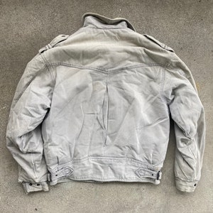 Puede incluir: Una chaqueta utilitaria gris claro con cierre de cremallera y cuello alto. La chaqueta tiene dos bolsillos con solapa en el pecho y dos bolsillos en la cintura.