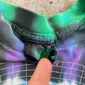 Puede incluir: Primer plano del cuello de una camiseta tie-dye con una combinaci&oacute;n de colores verde, azul y morado. La camiseta tiene una etiqueta negra cosida en el cuello.