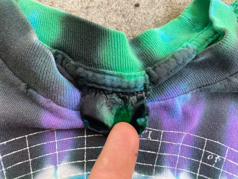 Puede incluir: Primer plano del cuello de una camiseta tie-dye con una combinaci&oacute;n de colores verde, azul y morado. La camiseta tiene una etiqueta negra cosida en el cuello.