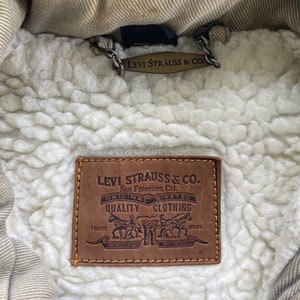 Puede incluir: Un parche de cuero marr&oacute;n Levi Strauss & Co. con el texto "Levi Strauss & Co. San Francisco, Cal. Original Riveted Quality Clothing Trade Mark" en letras blancas.
