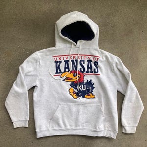 以下が含まれることがあります： グレーのフード付きスウェットシャツに「University of Kansas」の文字と、青と黄色のジャージを着た漫画のキャラクターが描かれています。キャラクターは、赤いクチバシと黄色い足を持つ鳥です。