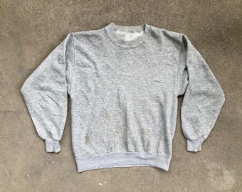 May include: A heather gray sweatshirt with a crew neck and long sleeves. The cuffs and hem are ribbed. The garment has some discoloration and wear, suggesting it is vintage or pre-owned. The sweatshirt is laid flat on a concrete surface.