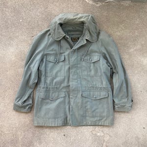 May include: A light green jacket with a large collar and four front pockets. The jacket has a drawstring waist and buttoned cuffs. The fabric appears to be a durable, possibly military-style material. The jacket is laid flat on a concrete surface.