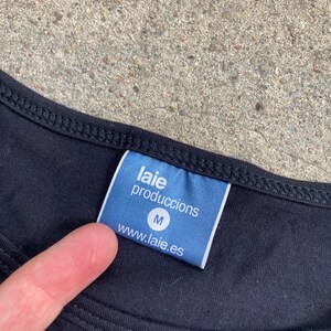 Puede incluir: Camiseta negra con una etiqueta azul que dice "laie produccions" y "www.laie.es". La etiqueta tambi&eacute;n tiene la letra "M". La camiseta est&aacute; sobre una superficie gris.