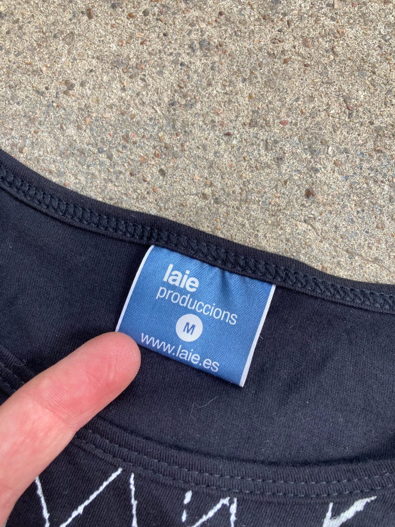Puede incluir: Camiseta negra con una etiqueta azul que dice "laie produccions" y "www.laie.es". La etiqueta tambi&eacute;n tiene la letra "M". La camiseta est&aacute; sobre una superficie gris.