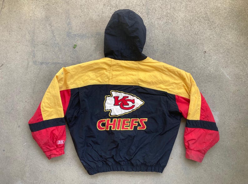 May include: A vintage Kansas City Chiefs hooded jacket. The jacket features a black hood and a color block design with yellow, red, and navy blue panels. The Chiefs logo and team name are prominently displayed on the back.