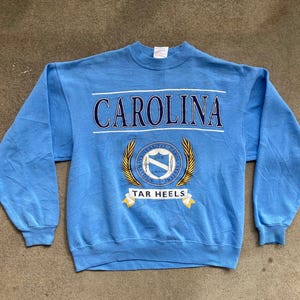 May include: A light blue sweatshirt with the text "CAROLINA" in navy blue letters and a crest with the text "UNIVERSITY OF NORTH CAROLINA" and "TAR HEELS" in gold letters.
