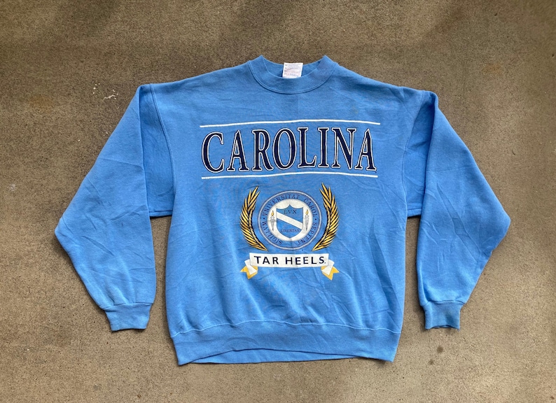 May include: A light blue sweatshirt with the text "CAROLINA" in navy blue letters and a crest with the text "UNIVERSITY OF NORTH CAROLINA" and "TAR HEELS" in gold letters.