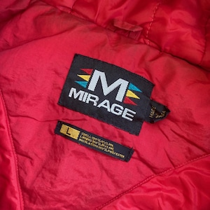 May include: Close-up of a red jacket interior with black labels. The main label reads "MIRAGE" with a stylized "M" and colorful accents. A smaller label indicates the size "L" and the materials: 100% nylon shell, lining, and insulation.