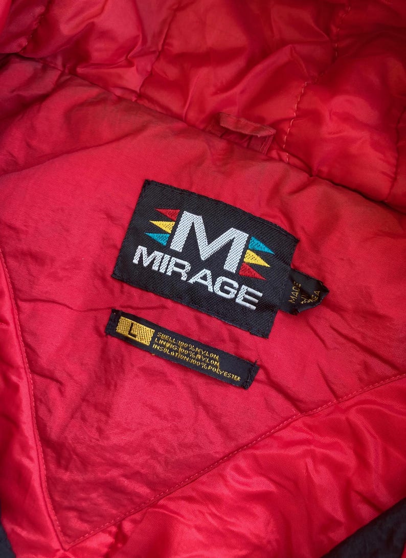 May include: Close-up of a red jacket interior with black labels. The main label reads "MIRAGE" with a stylized "M" and colorful accents. A smaller label indicates the size "L" and the materials: 100% nylon shell, lining, and insulation.