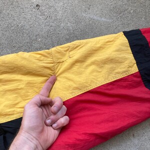 May include: Close-up of a section of a colorful jacket. The jacket features a color block design with yellow, red, and black panels. The fabric appears to be a lightweight, possibly nylon material. The stitching and seams are visible.